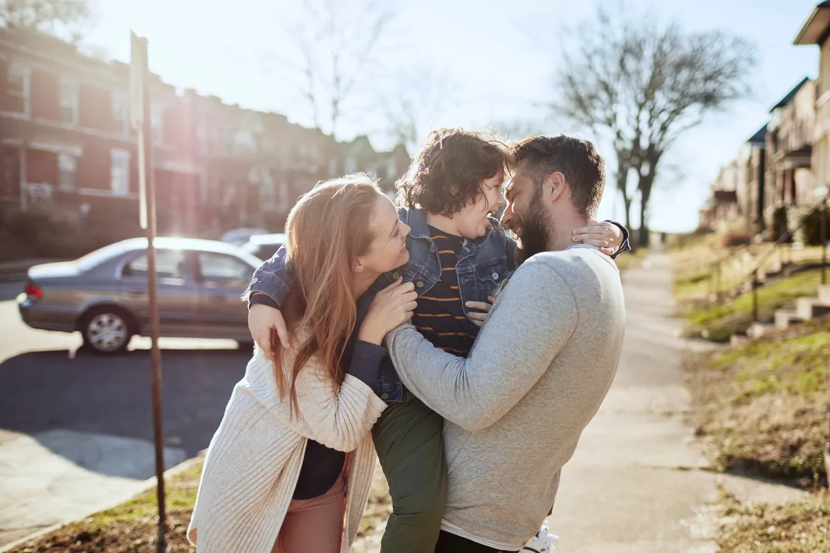 A mom and dad hugging their young son on a sunny sidewalk